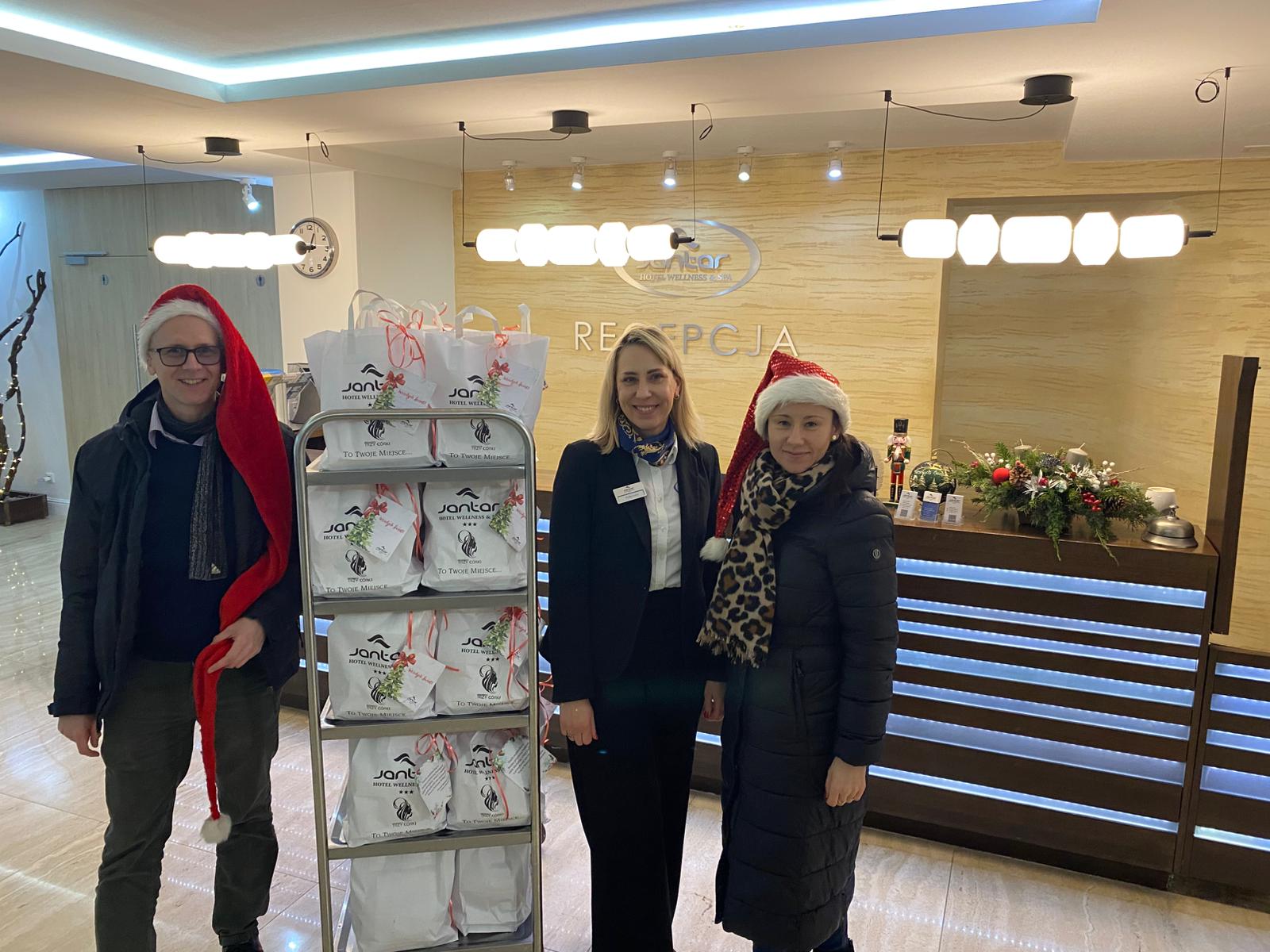 Recepcja hotelu wellness. Trzy osoby uśmiechają się do aparatu, stojąc obok wózka z torbami prezentowymi. Dwie osoby mają czapki Mikołaja. W tle widoczna lada recepcyjna i świąteczne dekoracje