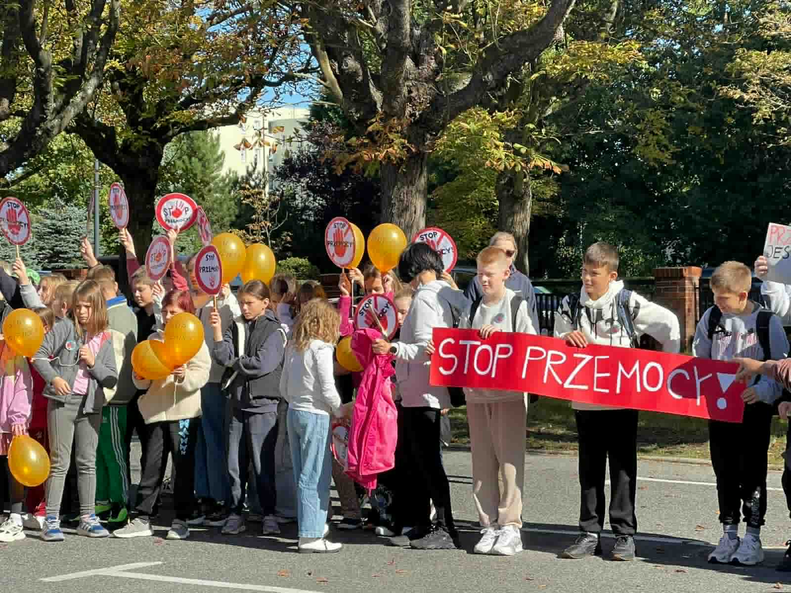 Grupa dzieci i młodzieży stoi na ulicy podczas marszu lub demonstracji przeciw przemocy. Uczestnicy trzymają pomarańczowe balony oraz plakaty z napisem 'STOP PRZEMOCY'. Na pierwszym planie widoczna jest czerwona tablica z napisem 'STOP PRZEMOCY'. W tle drzewa z jesiennymi liśćmi oraz budynki. Wydarzenie odbywa się w słoneczny dzień."
